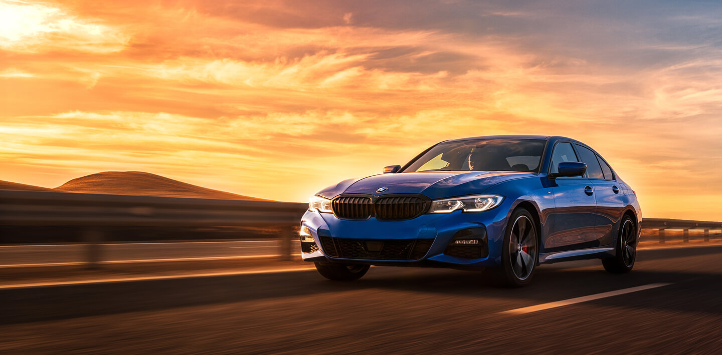 Blue BMW driving on highway at sunset with dramatic sky in the background.